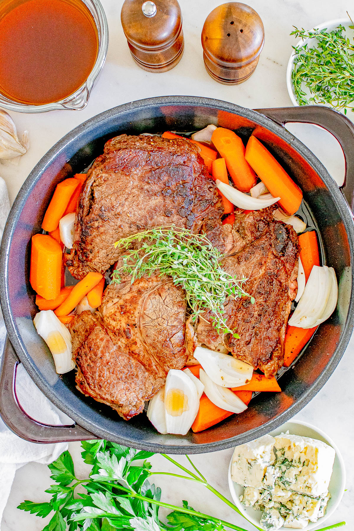 A pot roast with beef, carrots, onions, and herbs in a black pot, surrounded by broth, herbs, and cheese on a table.