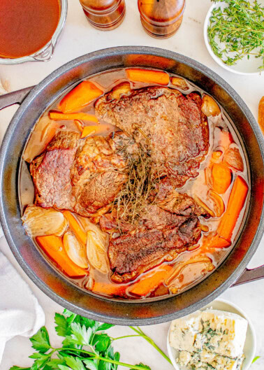 A stew pot with cooked beef, carrots, and onions, garnished with herbs, surrounded by ingredients like broth, herbs, and blue cheese on a white surface.