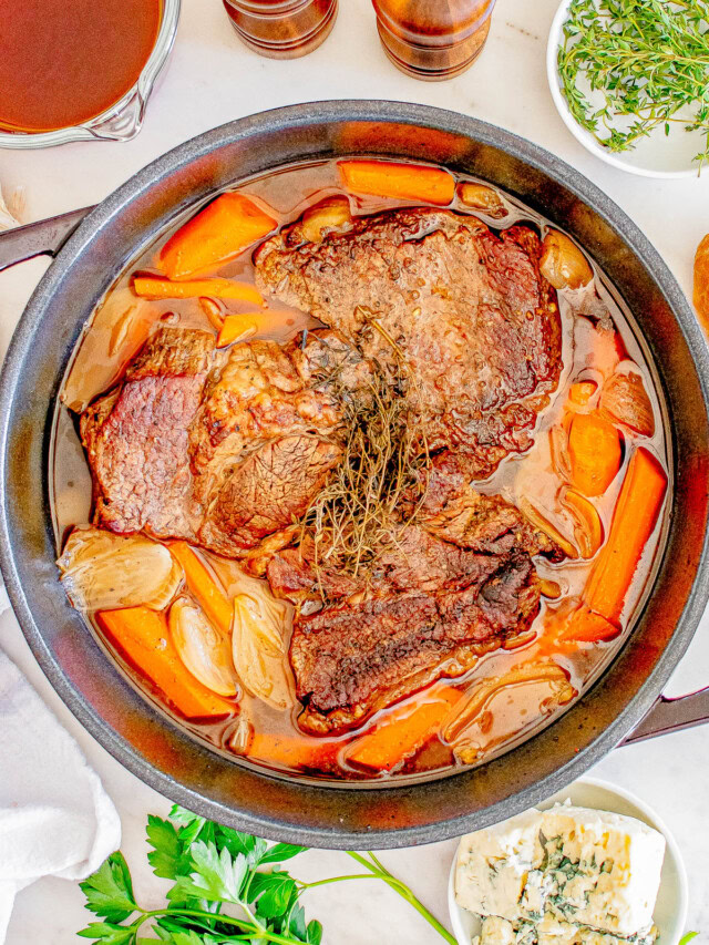 A stew pot with cooked beef, carrots, and onions, garnished with herbs, surrounded by ingredients like broth, herbs, and blue cheese on a white surface.