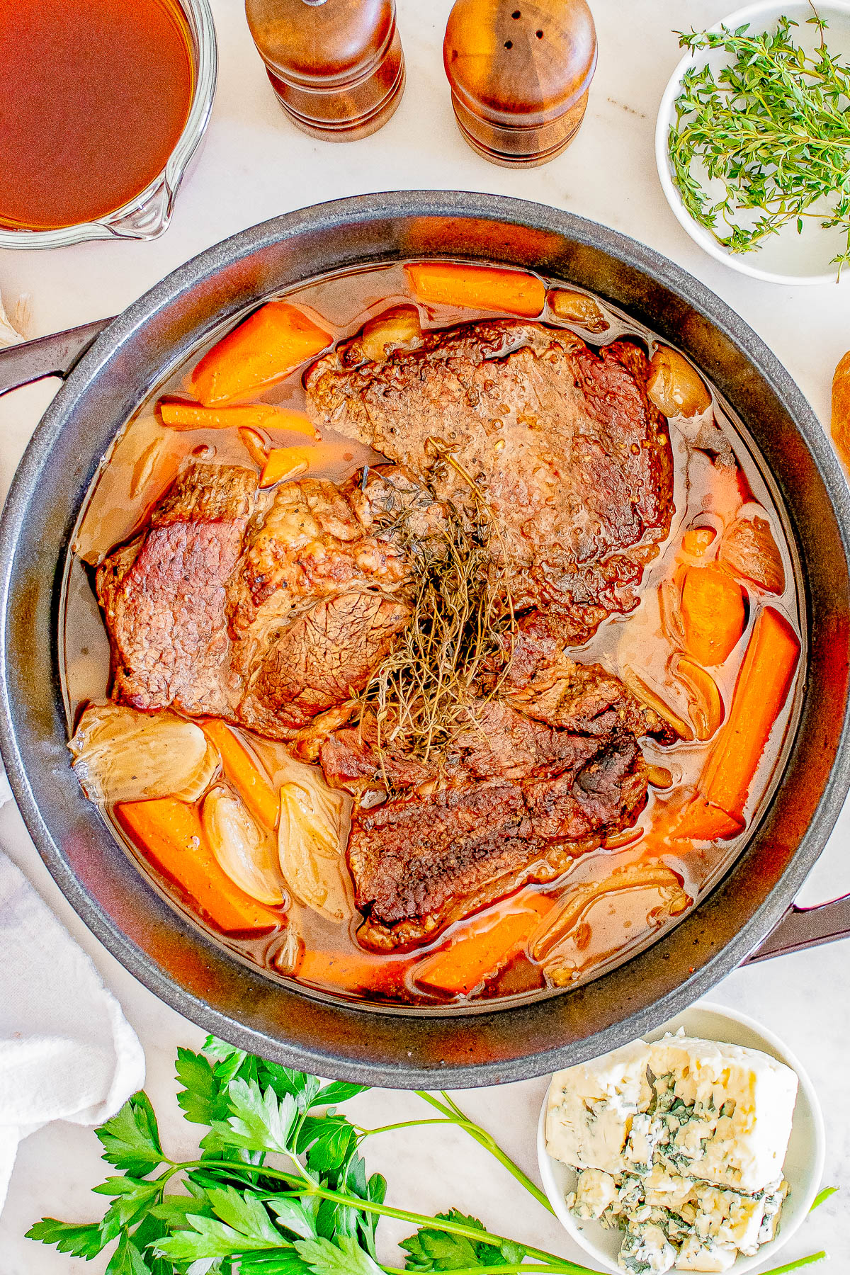 A stew pot with cooked beef, carrots, and onions, garnished with herbs, surrounded by ingredients like broth, herbs, and blue cheese on a white surface.