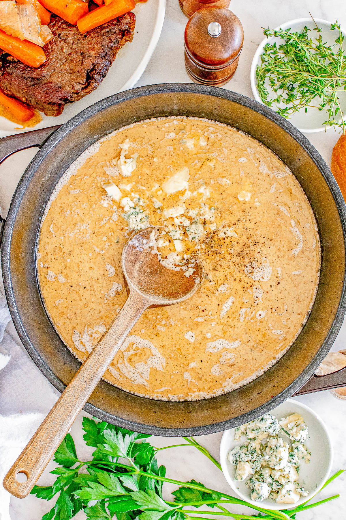 A pan filled with creamy sauce and crumbled cheese, surrounded by herbs, a dish of meat with carrots, and a wooden spoon resting inside the pan.