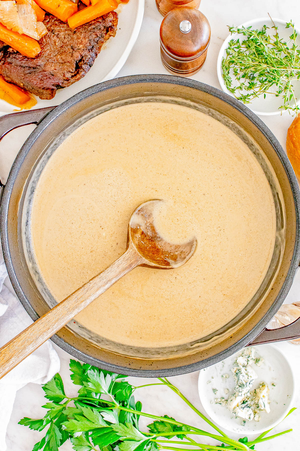 A pot of creamy gravy with a wooden ladle, surrounded by herbs, blue cheese, carrots, and beef on a plate.