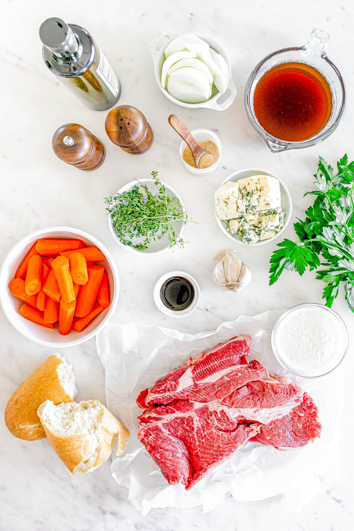 Assorted ingredients for a beef dish on a white surface, including raw beef, carrots, onions, herbs, broth, seasonings, bread, and cheese.