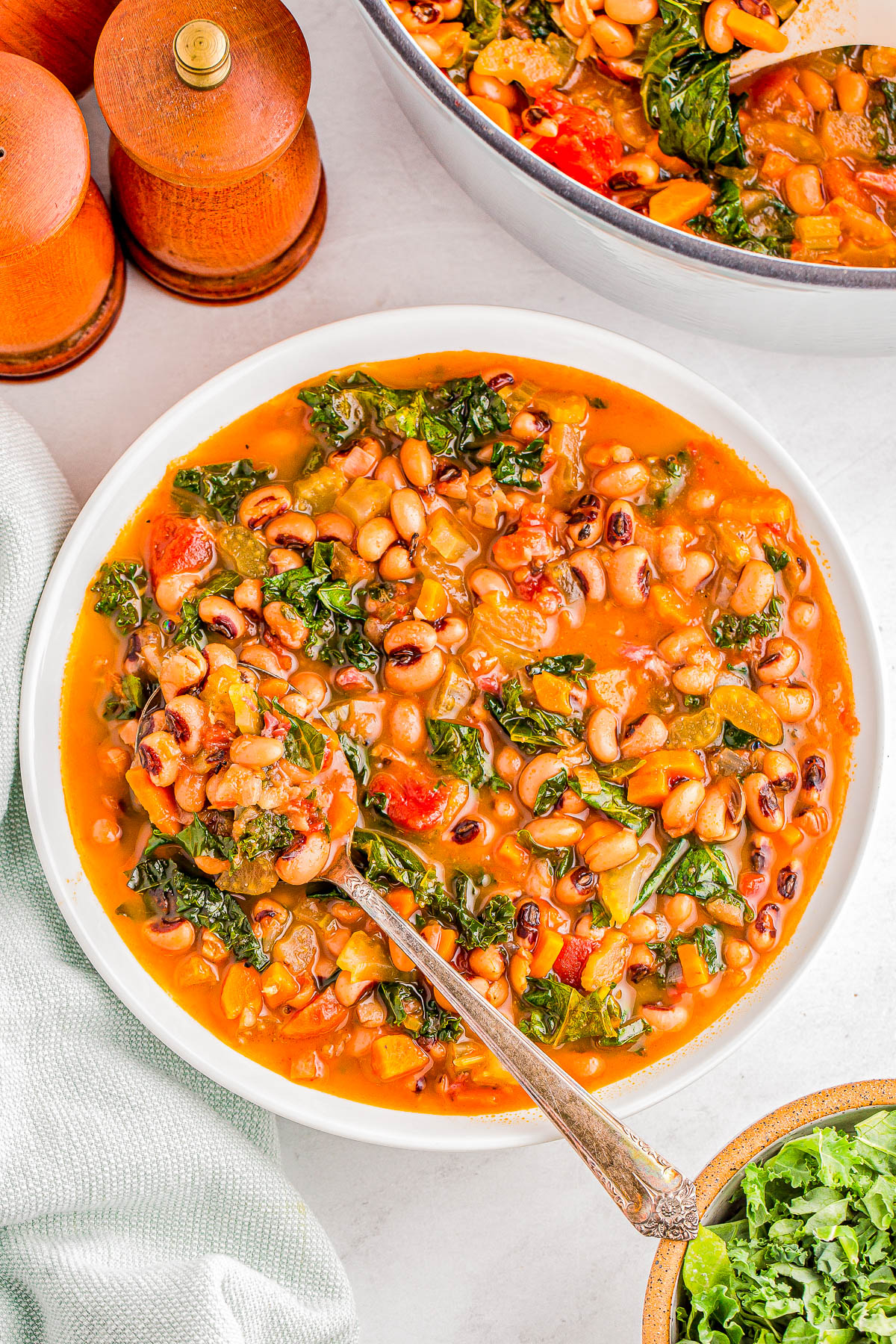 A bowl of vegetable soup with black-eyed peas, tomatoes, and greens on a white surface. A spoon rests in the bowl. Salt and pepper shakers are in the background.