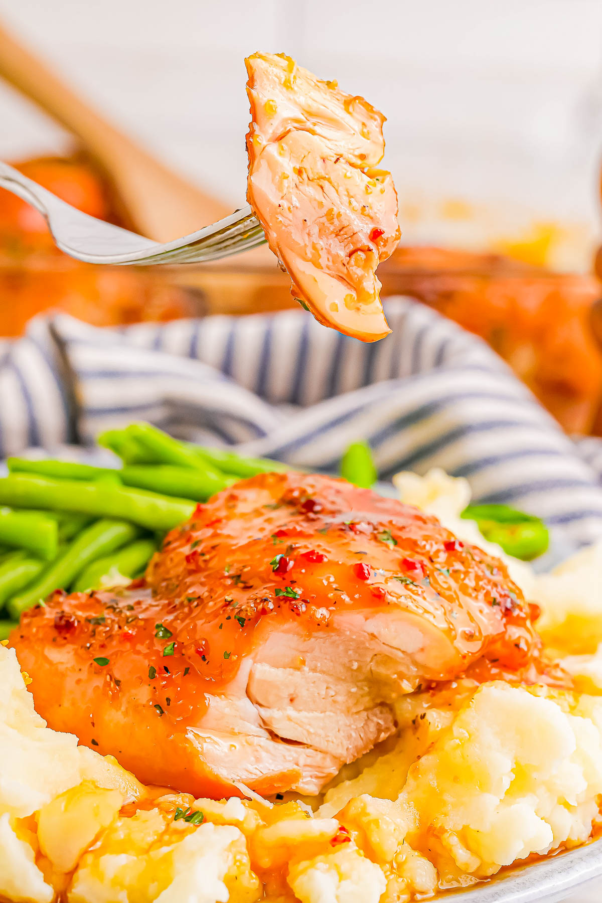 Close-up of a fork holding a piece of glazed chicken above a plate with chicken, mashed potatoes, and green beans.