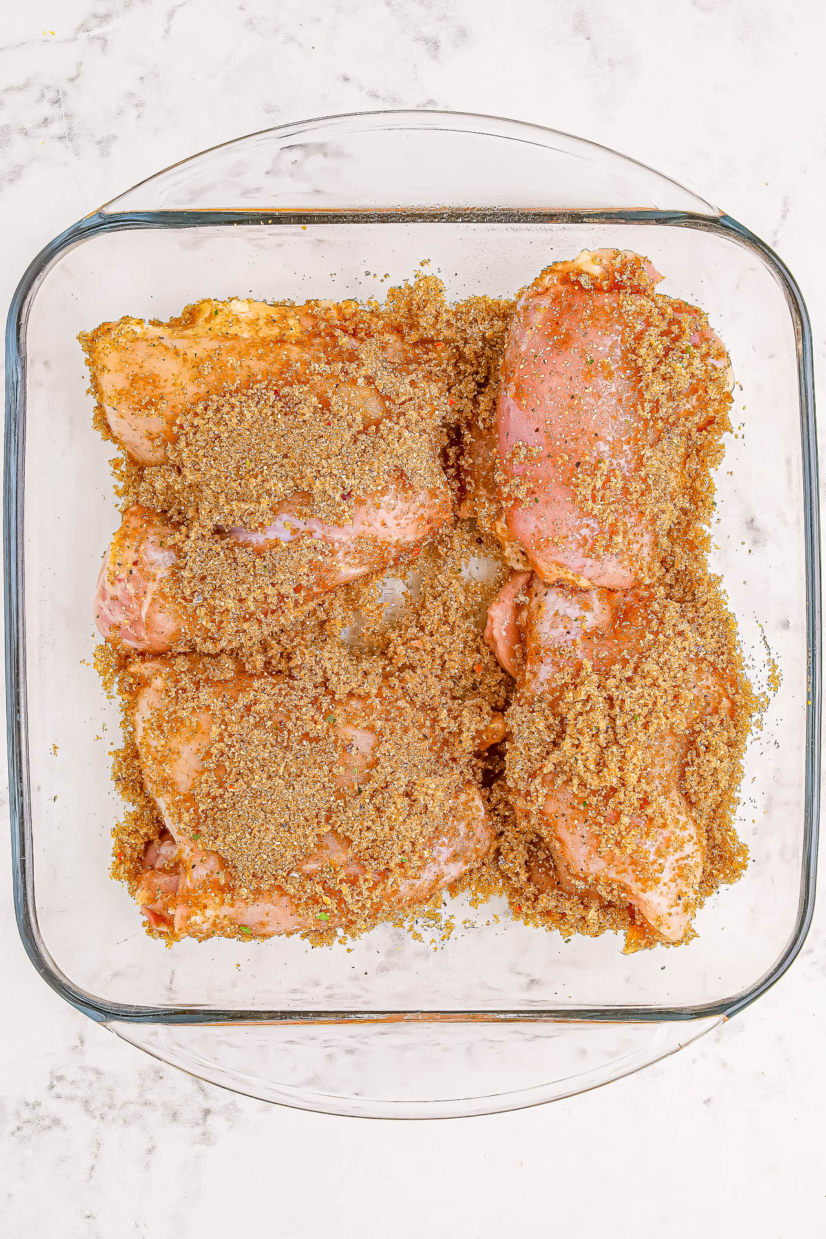 Raw chicken thighs seasoned with a brown sugar and spice mixture in a glass dish on a light marble countertop.