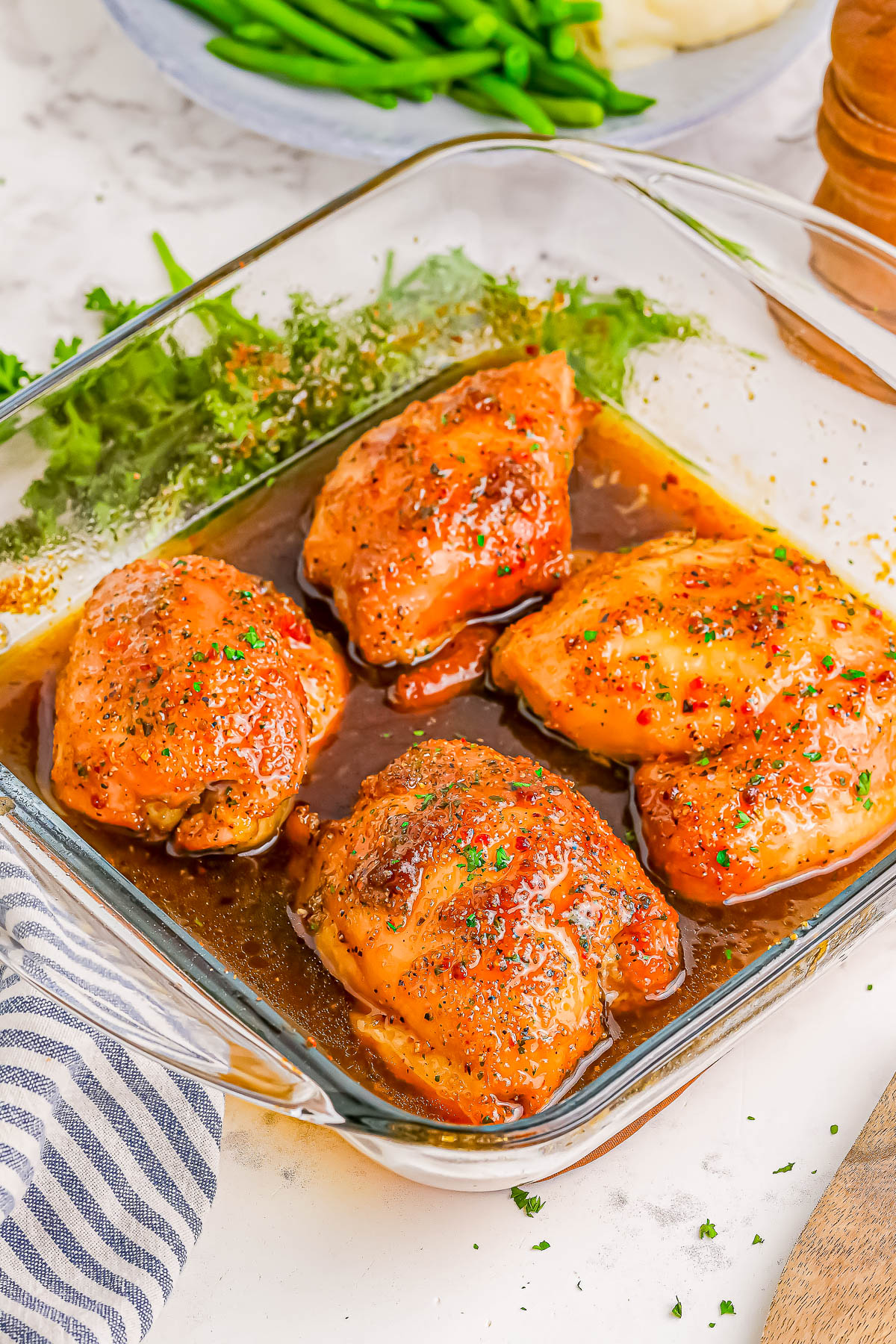 Four cooked chicken thighs in a glass baking dish, seasoned with sauce and herbs, on a kitchen counter with a striped towel and green beans in the background.