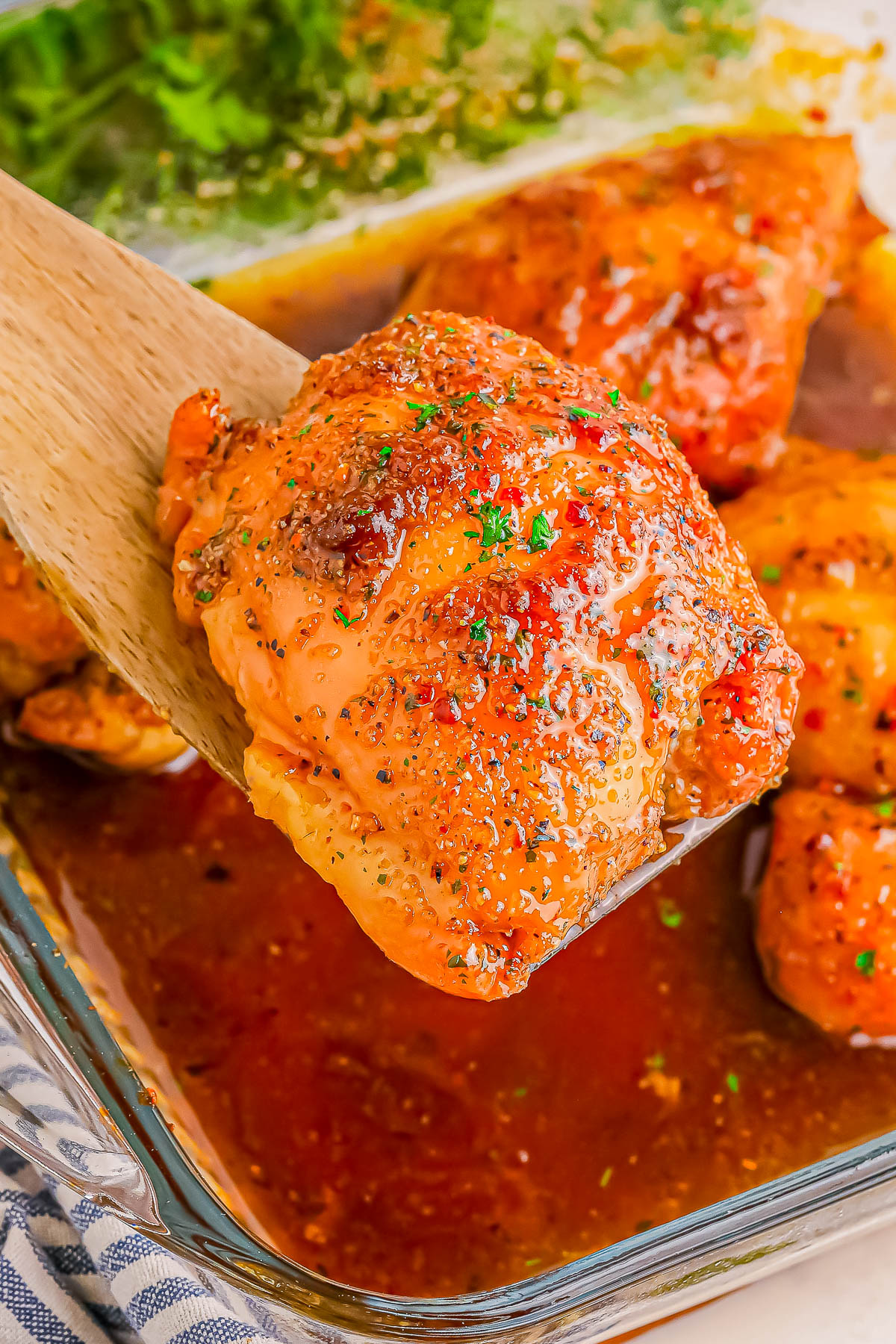 Close-up of a wooden spatula holding a glazed, baked chicken thigh. The background shows other chicken pieces in a glass baking dish with sauce.