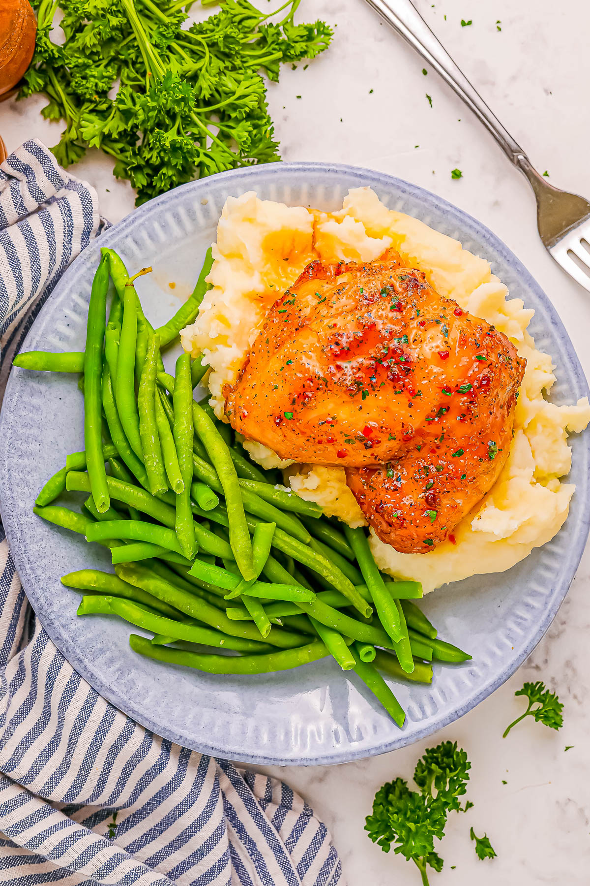 A plate with mashed potatoes, green beans, and a seasoned chicken breast on top. A striped napkin, fork, and parsley garnish are visible.