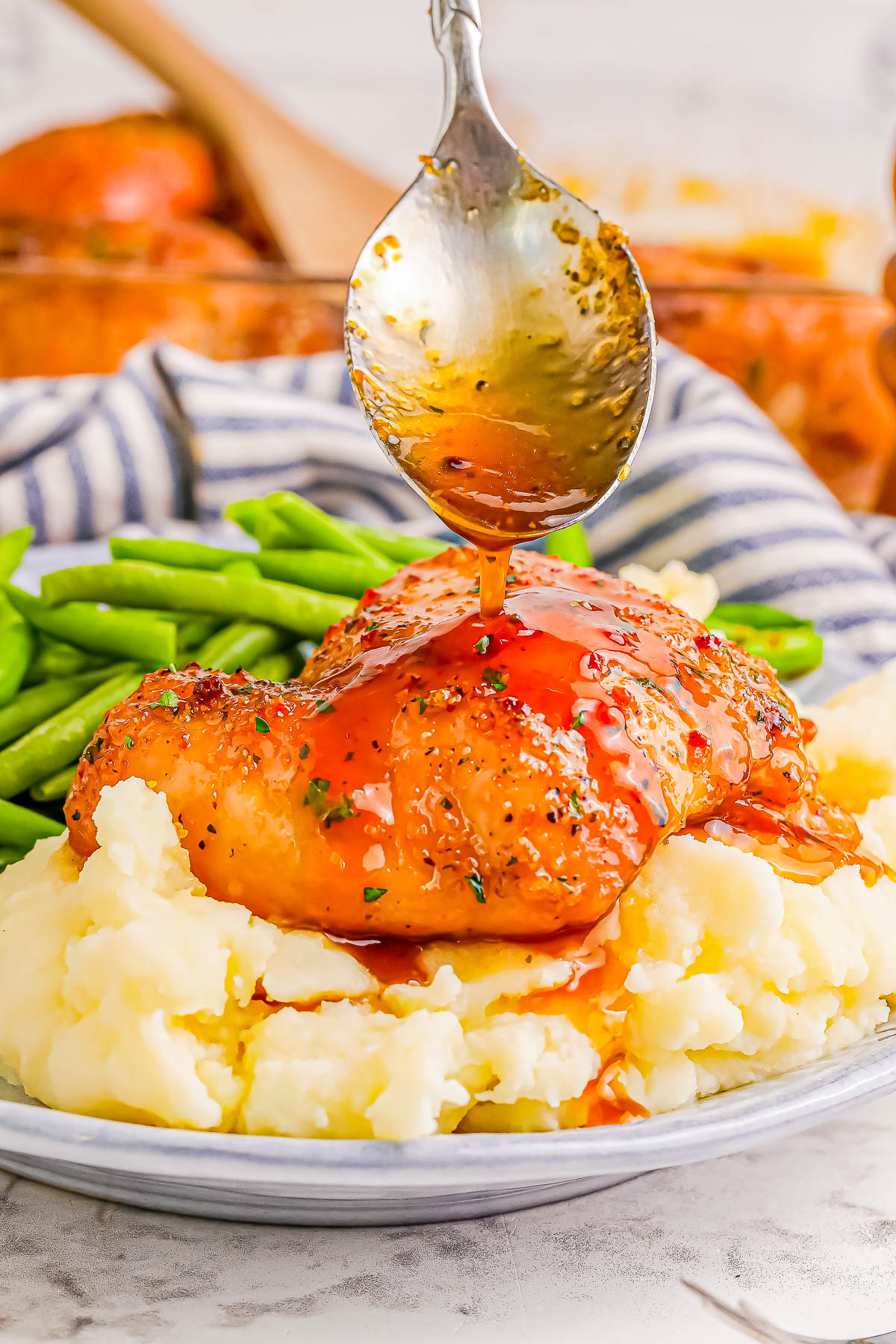 A spoon drizzles sauce over chicken served on mashed potatoes, accompanied by green beans.