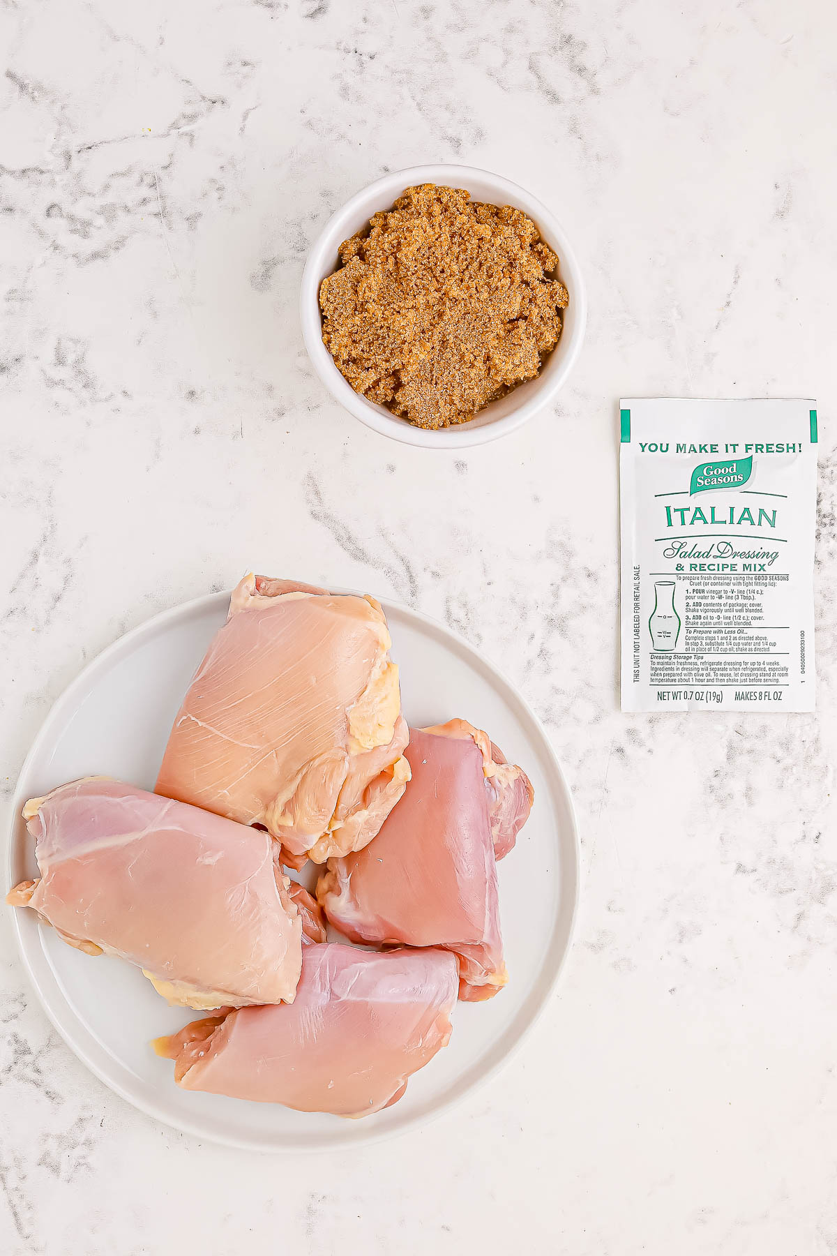 Raw chicken thighs on a plate, a bowl of mustard, and a packet of Italian dressing mix on a marble surface.