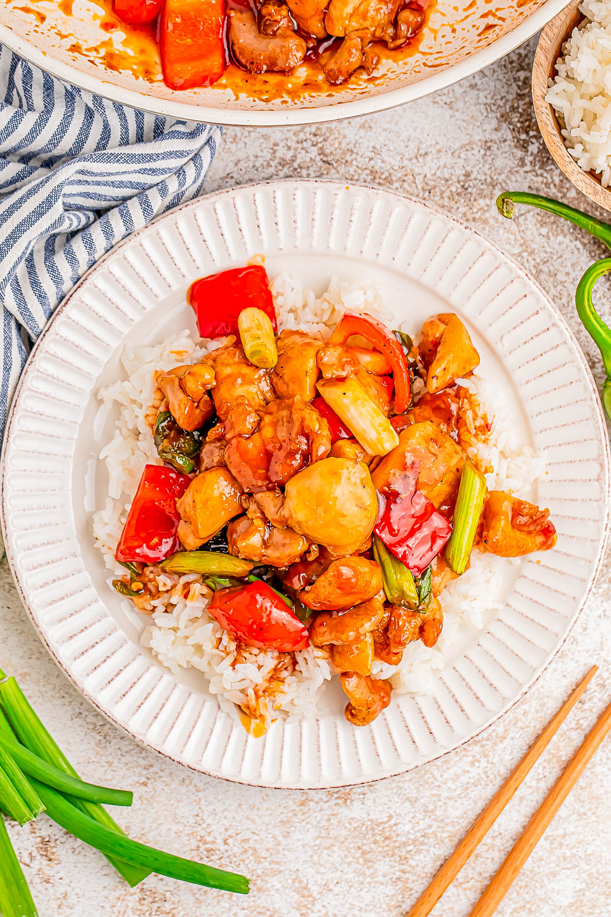 A plate of stir-fried chicken with bell peppers, spring onions, and sauce over white rice, accompanied by a pair of wooden chopsticks.