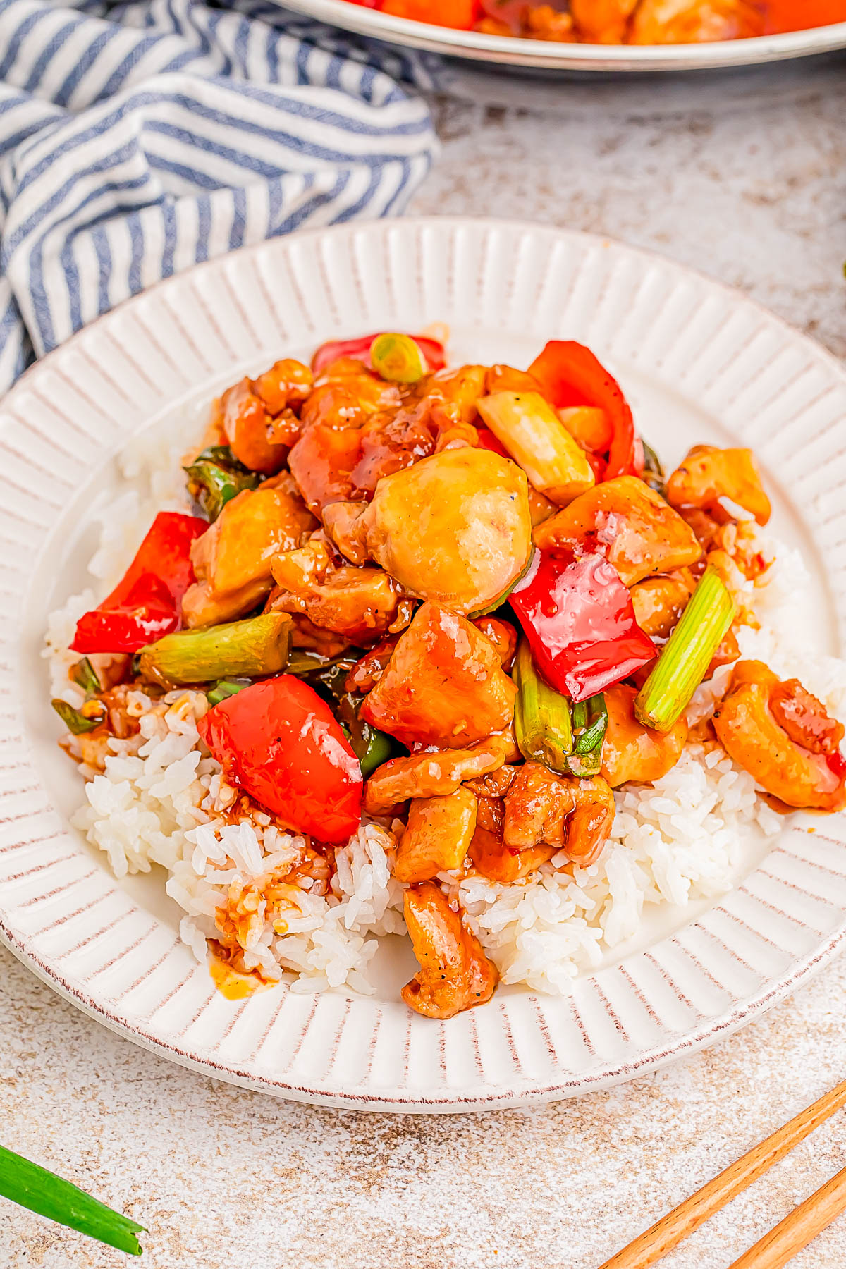 A plate of white rice topped with stir-fried chicken, red bell peppers, green vegetables, and sauce. A striped cloth napkin is visible in the background.