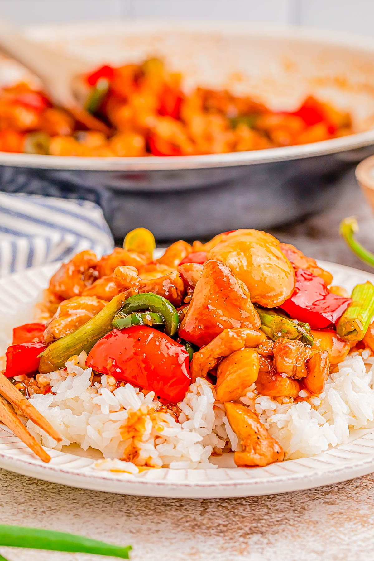 A plate of white rice topped with stir-fried chicken, red bell peppers, and green onions. A pan with more of the dish is visible in the background.