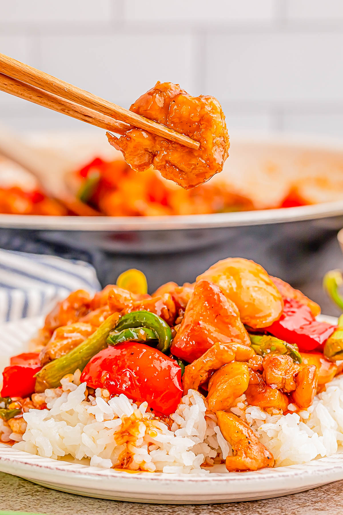 Chopsticks holding a piece of chicken above a plate of sweet and sour chicken with peppers and rice.