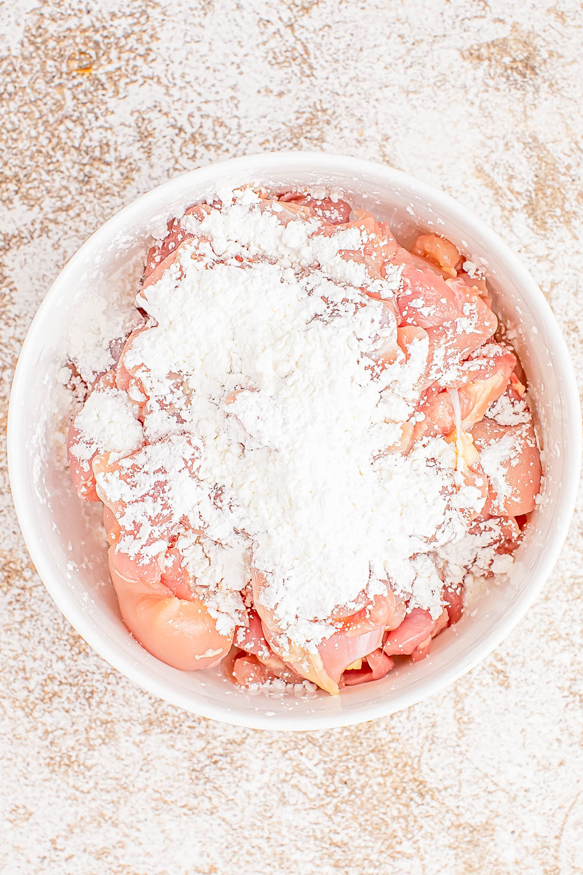 Raw chicken pieces in a bowl, covered with a powdery white seasoning on a textured surface.