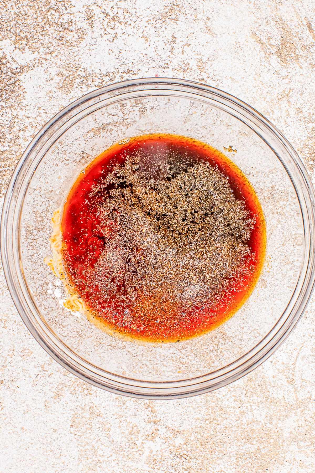 A glass bowl with a mixture of red liquid and black pepper on a speckled countertop.