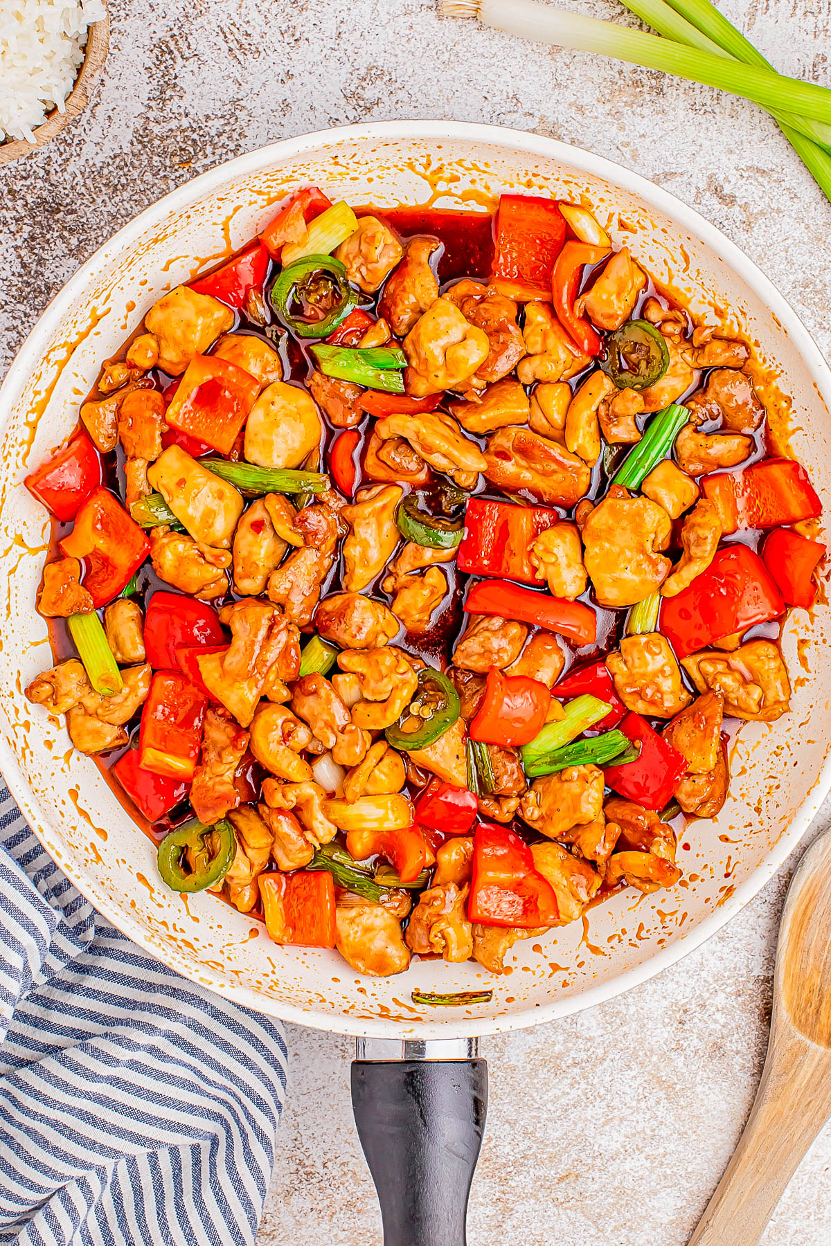 A pan of vibrant stir-fried chicken with red bell peppers, green onions, and a glazed sauce, placed on a rustic surface next to a striped cloth and wooden spoon.