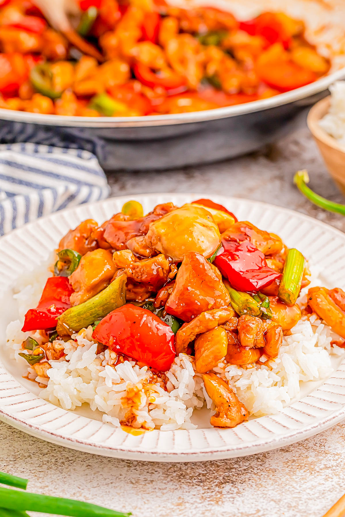 A plate of rice topped with stir-fried chicken, red peppers, and green onions, with a pan of more stir-fry in the background.