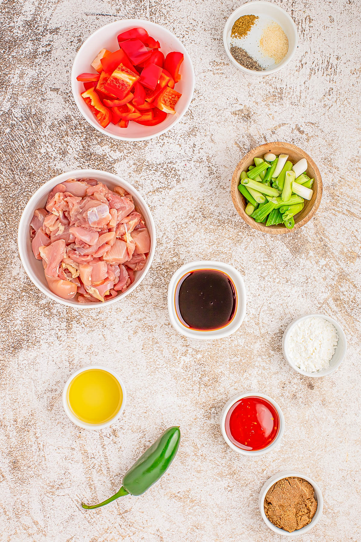 Ingredients arranged on a textured surface, including chopped chicken, red peppers, green onions, sauces, spices, a jalapeño, and seasonings in small bowls.