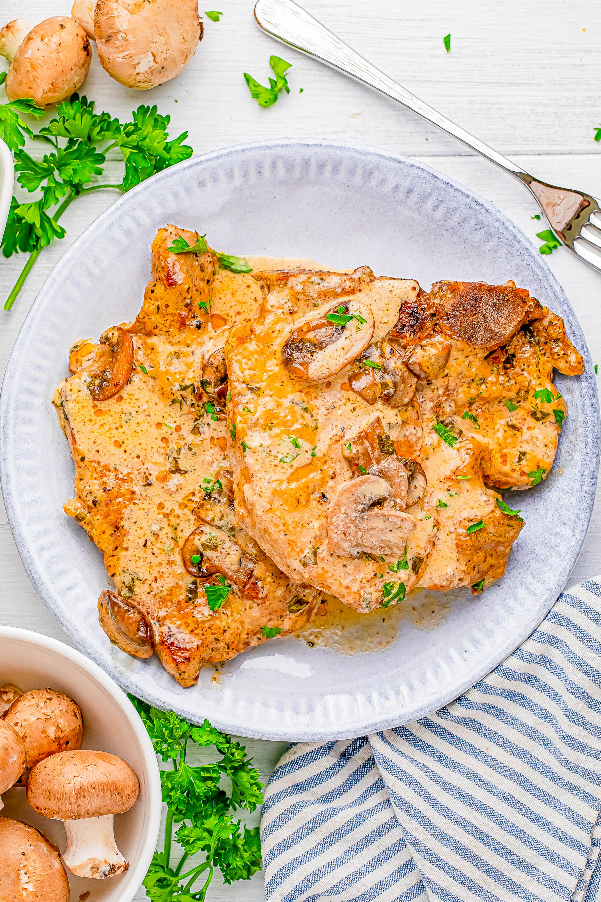 Plate of cooked pork chops topped with mushrooms and fresh herbs, with parsley and mushrooms on the side.