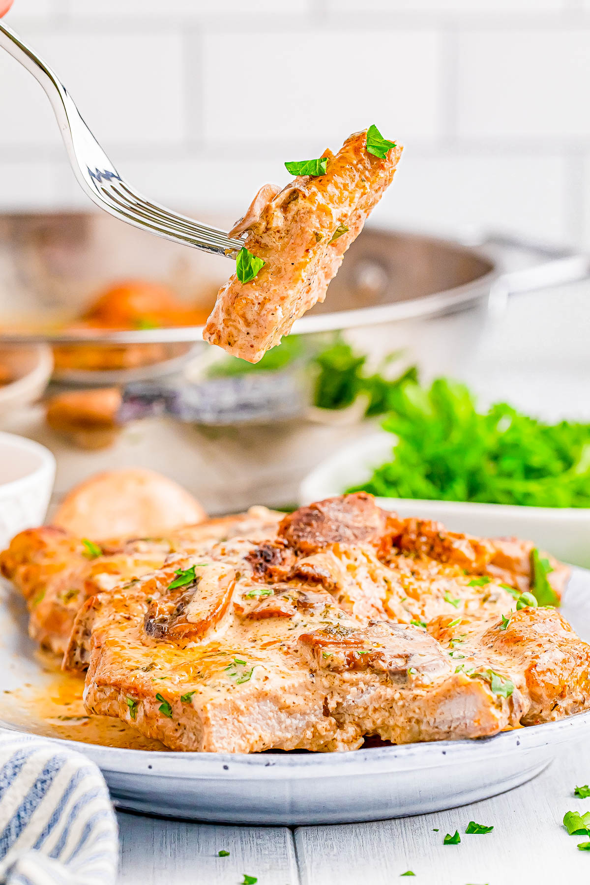 A fork holding a piece of seasoned cooked chicken over a plate of chicken with sauce. Fresh herbs are visible on top, with a sprig of parsley in the background.