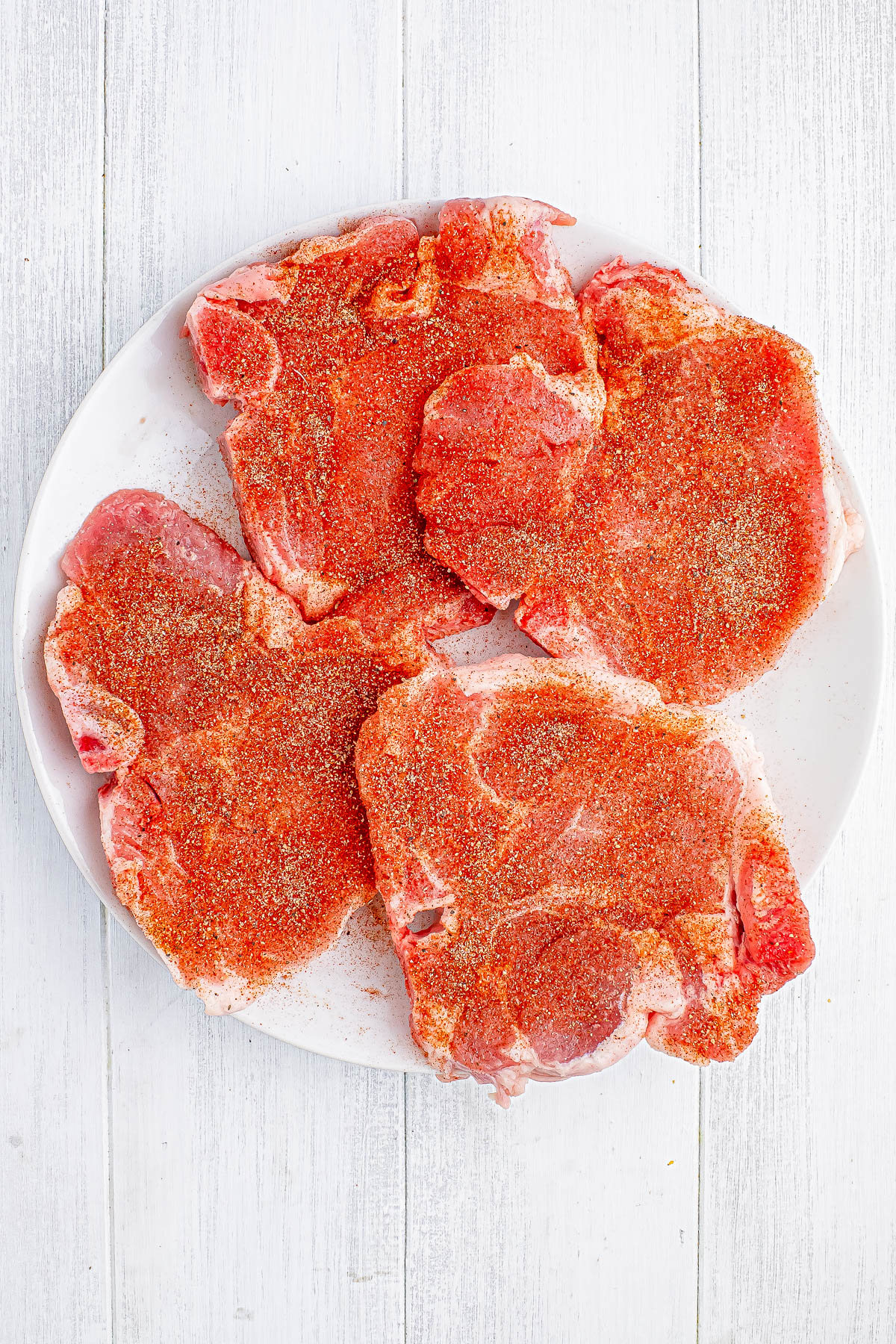 A plate of raw, seasoned pork chops on a white wooden surface.