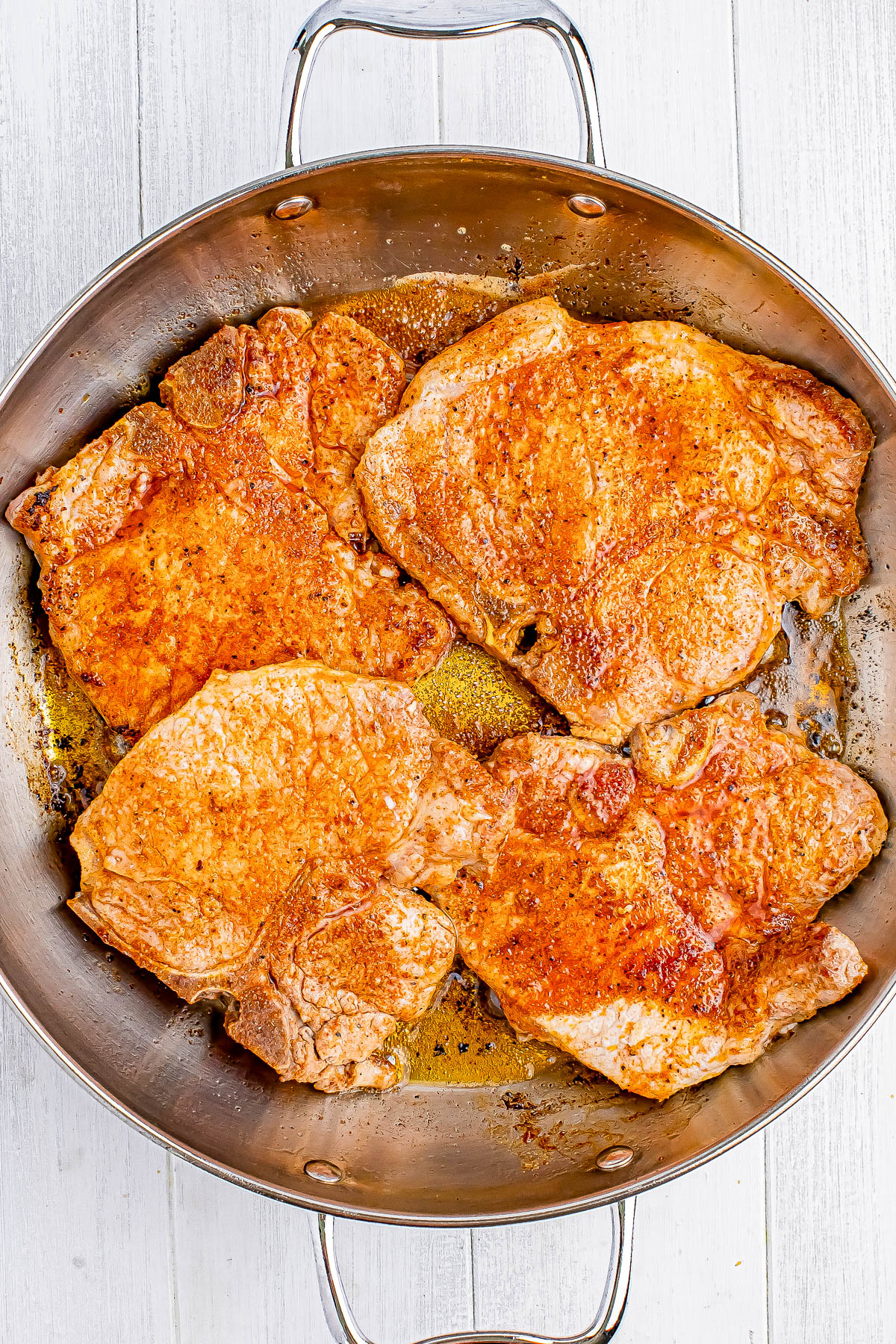 Four cooked pork chops in a metal frying pan on a white wooden surface.