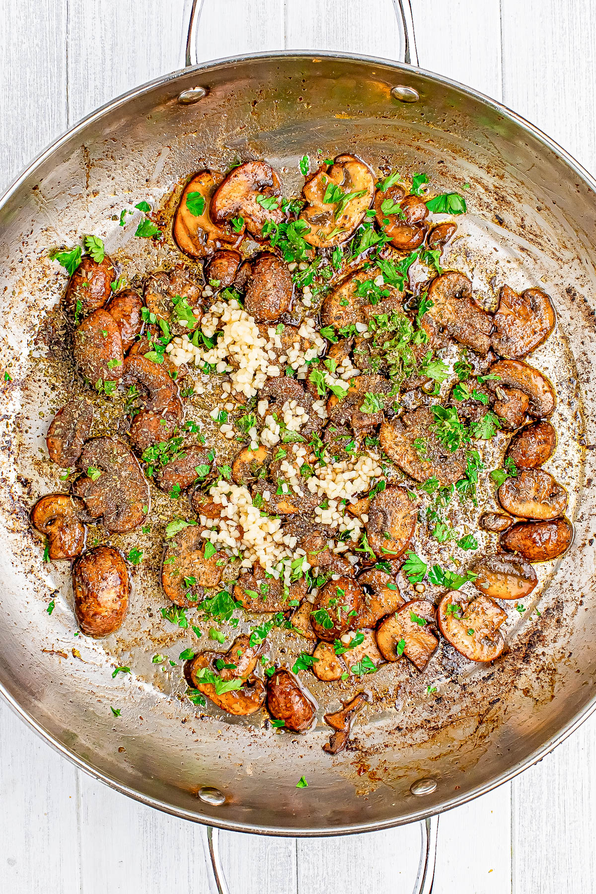 Sautéed mushrooms in a skillet with herbs and chopped garlic on a light wooden surface.