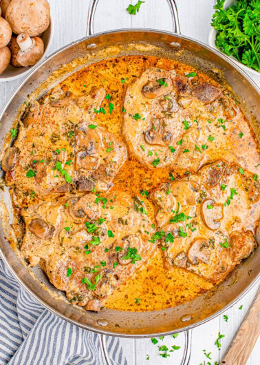 A pan containing cooked pork chops in a creamy mushroom sauce, garnished with chopped parsley. A bowl of mushrooms and parsley are in the background.