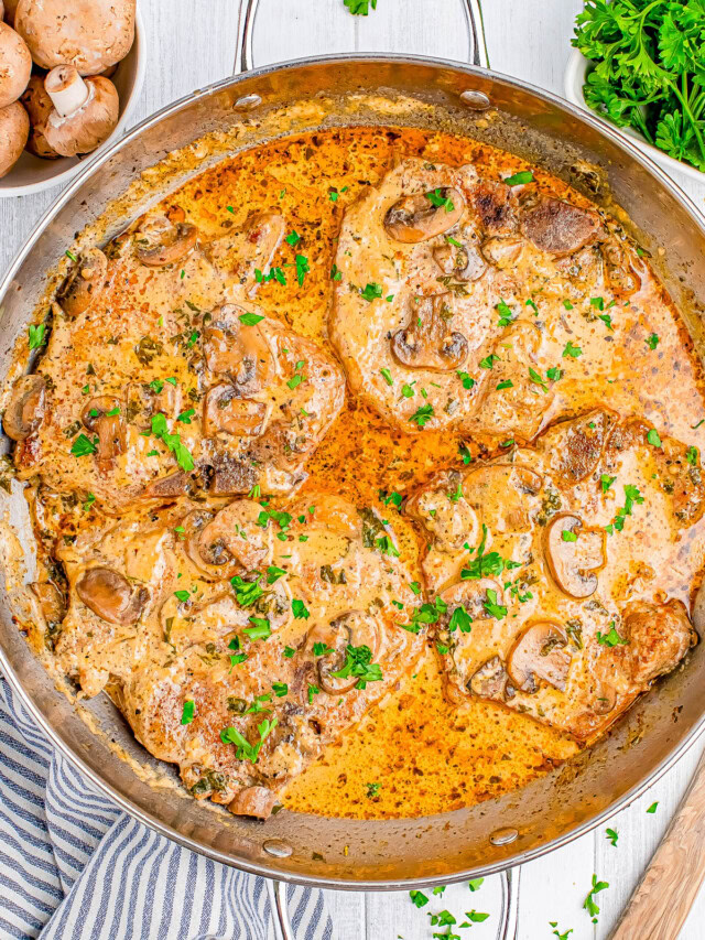 A pan containing cooked pork chops in a creamy mushroom sauce, garnished with chopped parsley. A bowl of mushrooms and parsley are in the background.