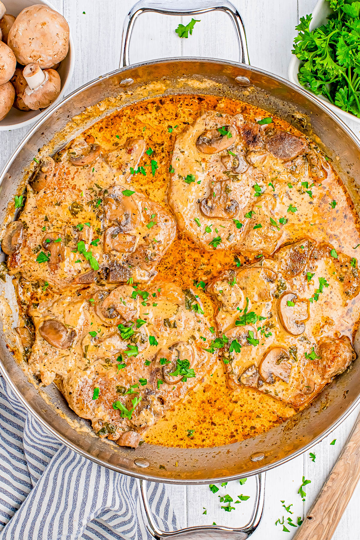 A pan containing cooked pork chops in a creamy mushroom sauce, garnished with chopped parsley. A bowl of mushrooms and parsley are in the background.