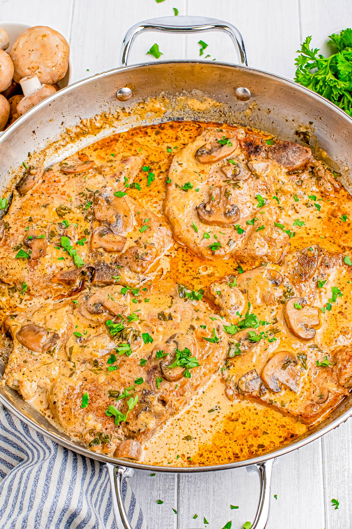 Pork chops in creamy mushroom sauce garnished with parsley in a skillet, with mushrooms and parsley on the side.