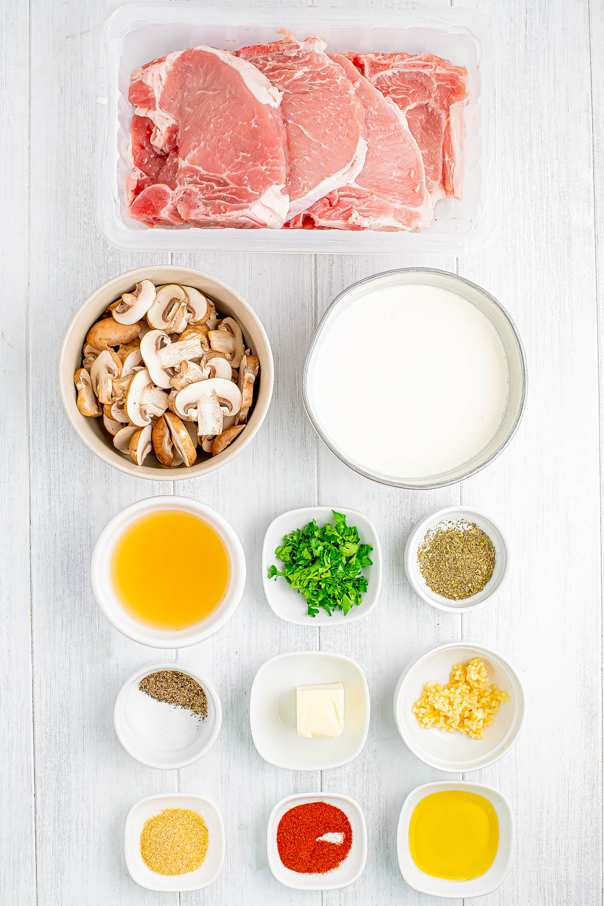 Top-down view of ingredients including raw pork chops, mushrooms, cream, broth, herbs, seasonings, butter, and garlic on a white surface.