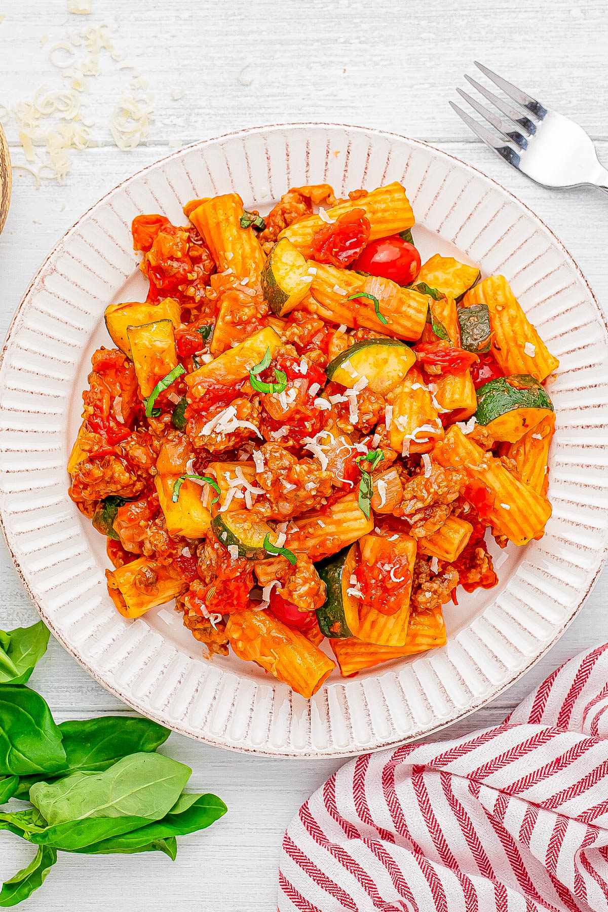 A plate of rigatoni pasta with tomato sauce, zucchini, and sprinkled cheese, served on a white plate. A fork, basil leaves, and a striped napkin are nearby.