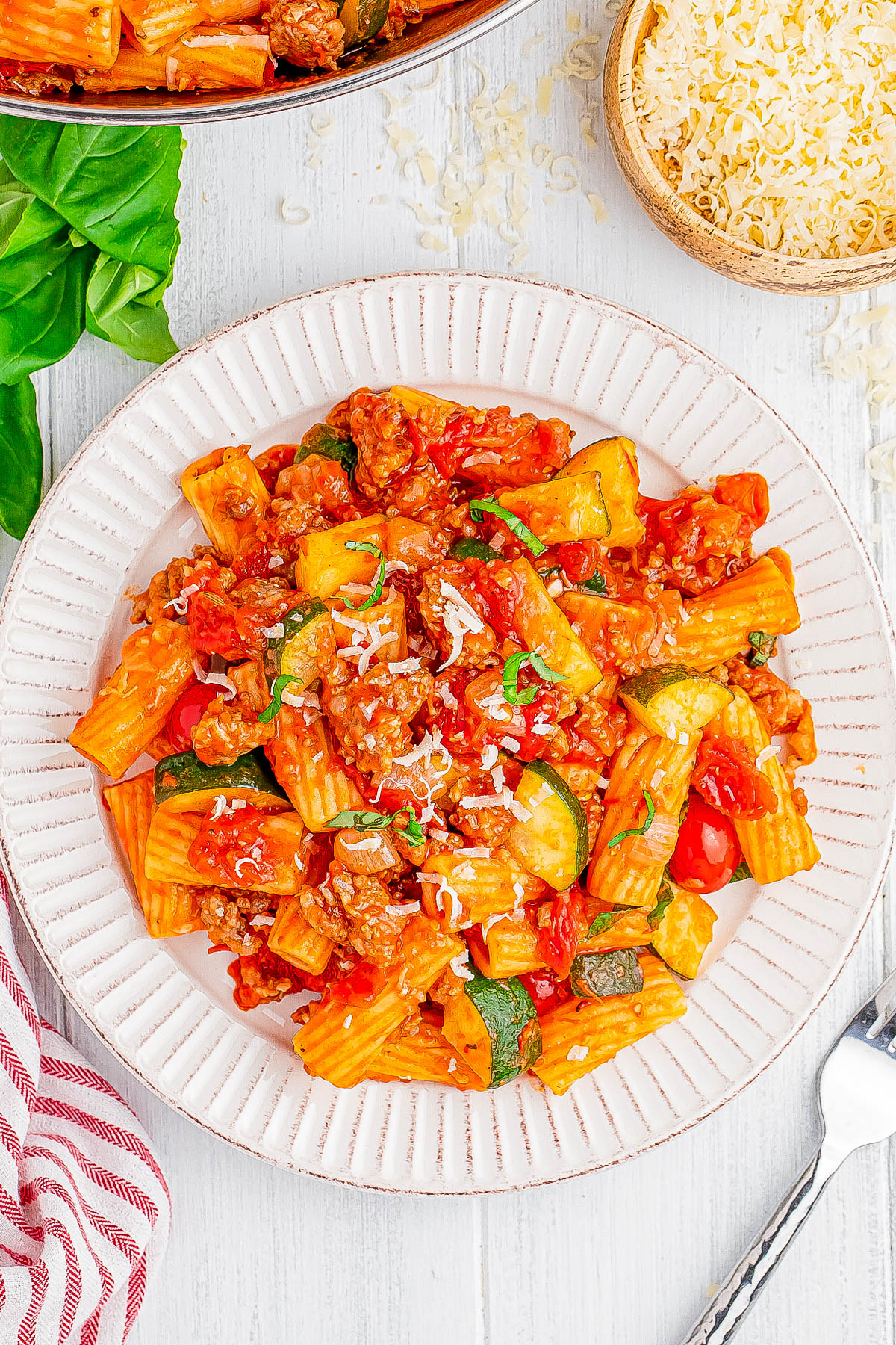 A plate of rigatoni pasta with red sauce, zucchini, and ground meat, garnished with cheese and herbs, on a white plate. A fork, basil, and grated cheese are visible nearby.