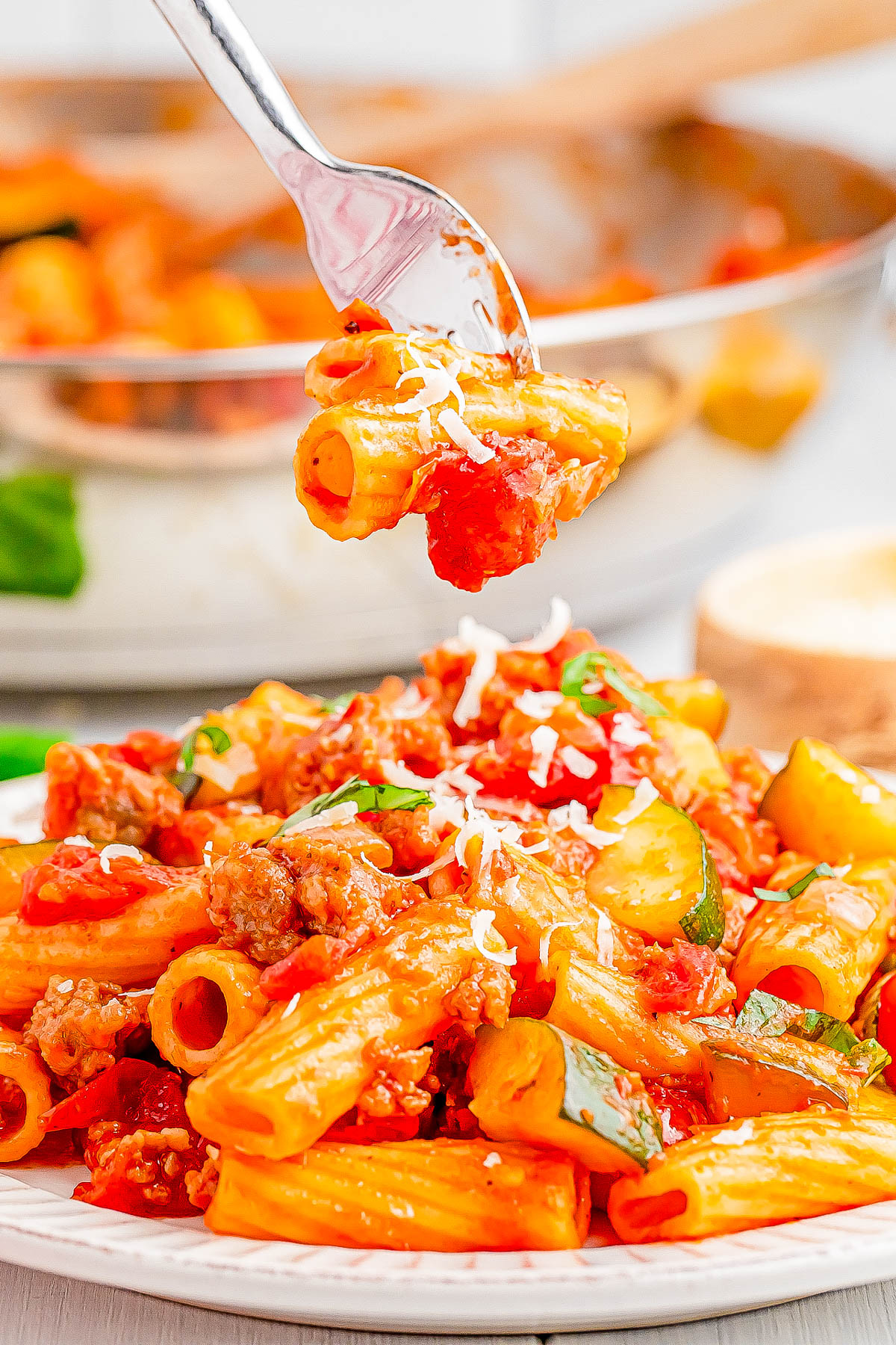 Plate of rigatoni with tomato sauce, zucchini, and a sprinkle of cheese. A fork holds a portion above the plate. A pan with more pasta is in the background.