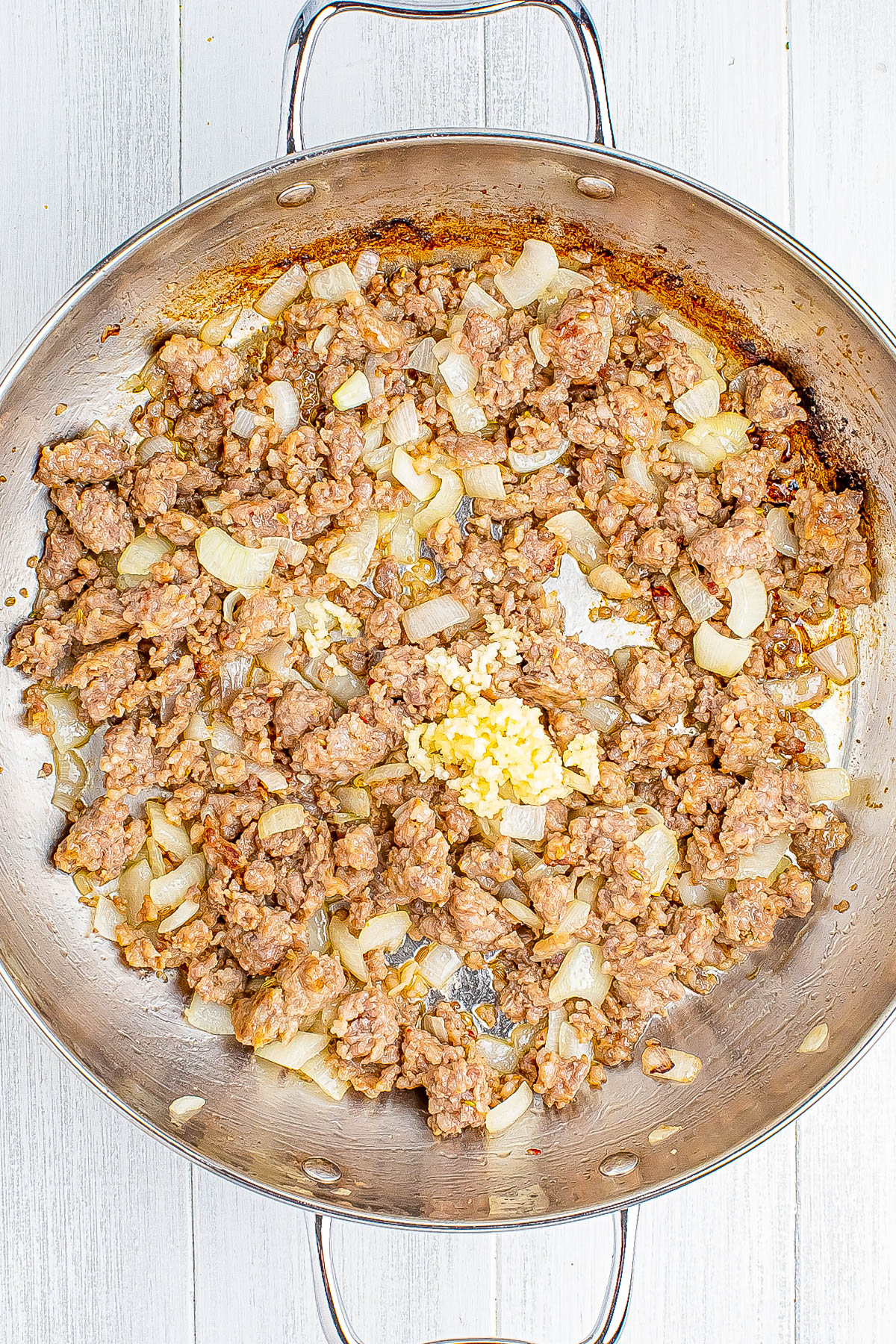 A skillet of browned ground meat with chopped onions and minced garlic on a light wood surface.