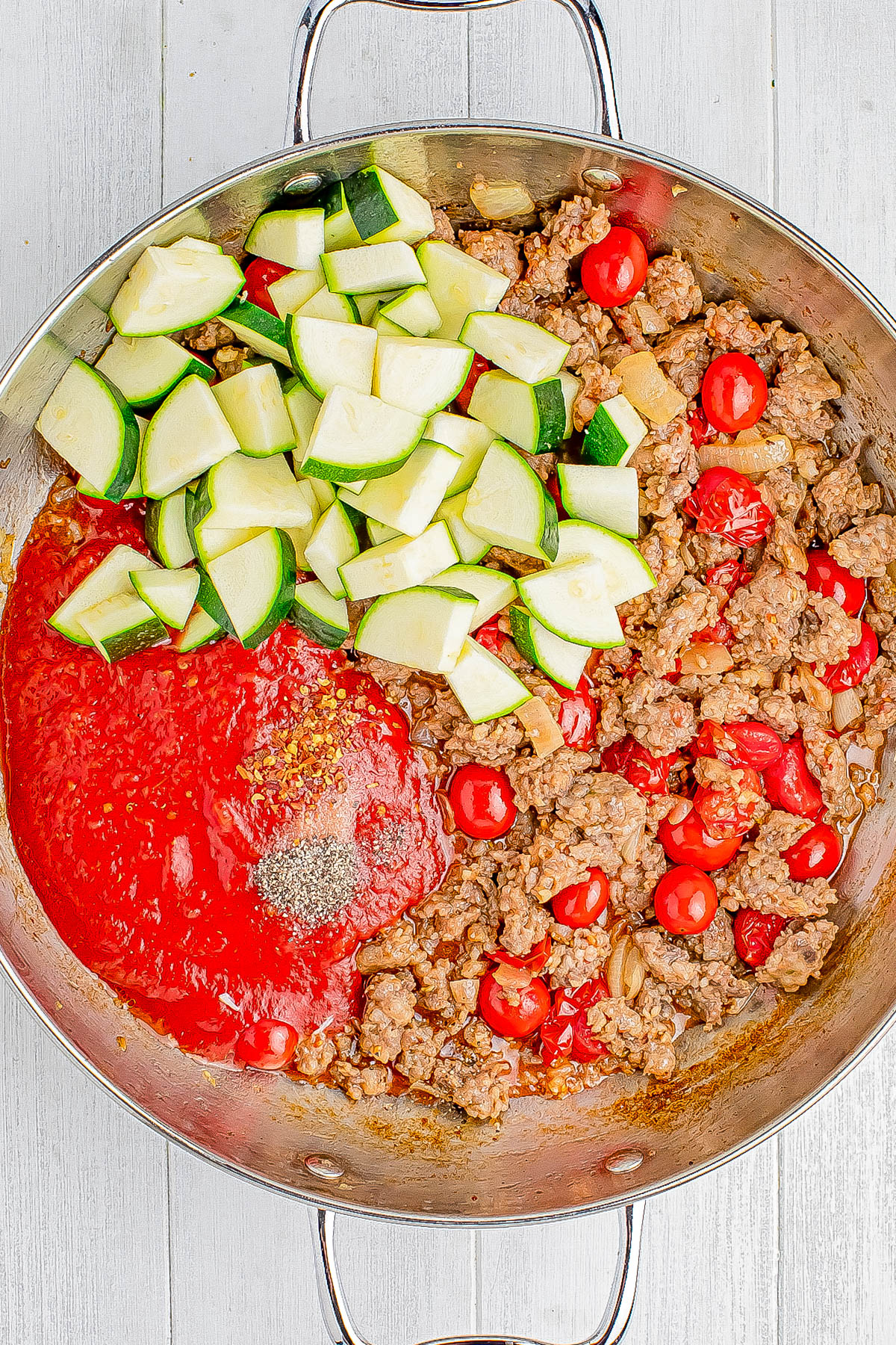 A skillet with ground meat, chopped zucchini, cherry tomatoes, and tomato sauce mixture, partially combined, on a white wooden surface.