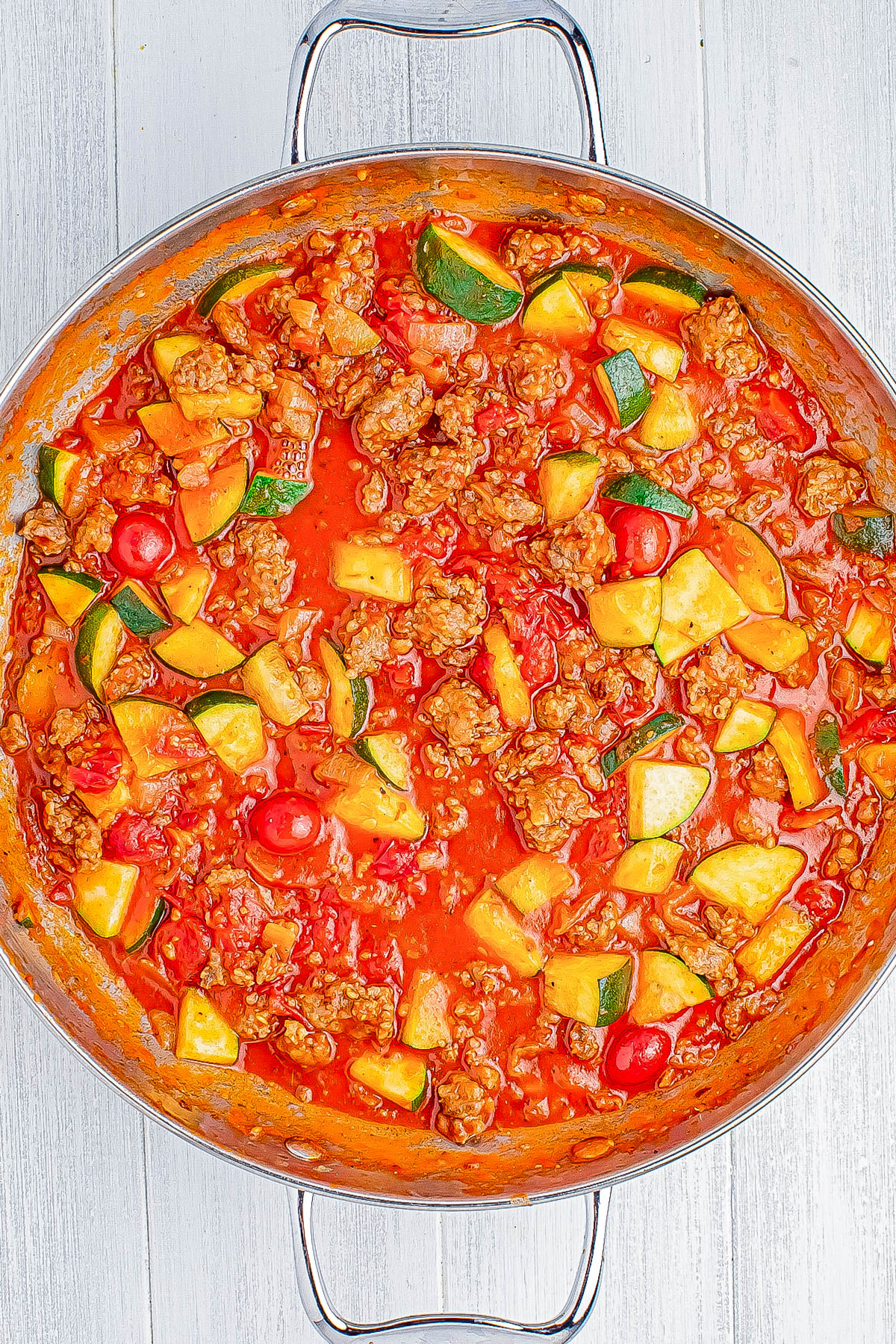 A skillet filled with a tomato-based stew containing sausage chunks, sliced zucchini, diced potatoes, and cherry tomatoes.