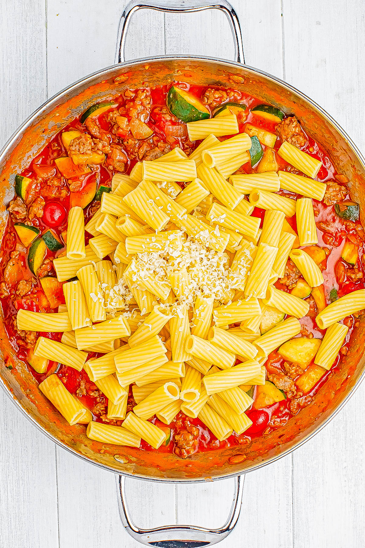 A pan of rigatoni pasta with zucchini, cherry tomatoes, and ground meat in a tomato sauce, topped with grated cheese, on a light wooden surface.