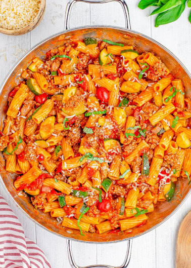 A pan of rigatoni pasta with ground meat, zucchini, cherry tomatoes, and grated cheese, garnished with fresh basil.