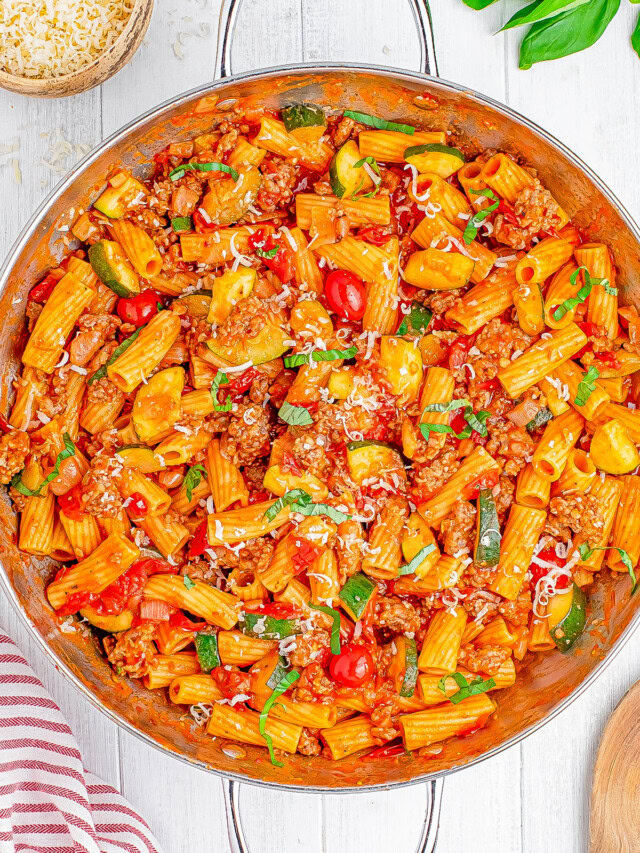 A pan of rigatoni pasta with ground meat, zucchini, cherry tomatoes, and grated cheese, garnished with fresh basil.