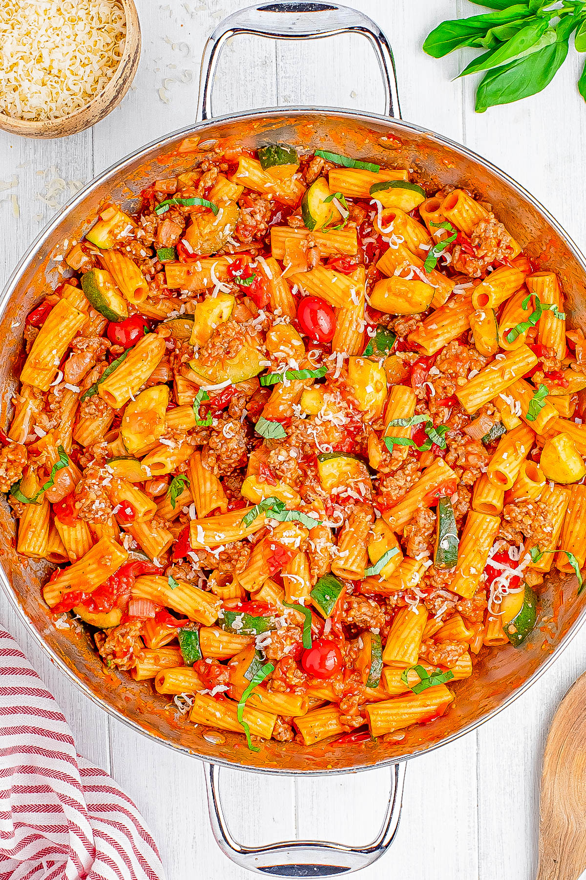 A pan of rigatoni pasta with ground meat, zucchini, cherry tomatoes, and grated cheese, garnished with fresh basil.