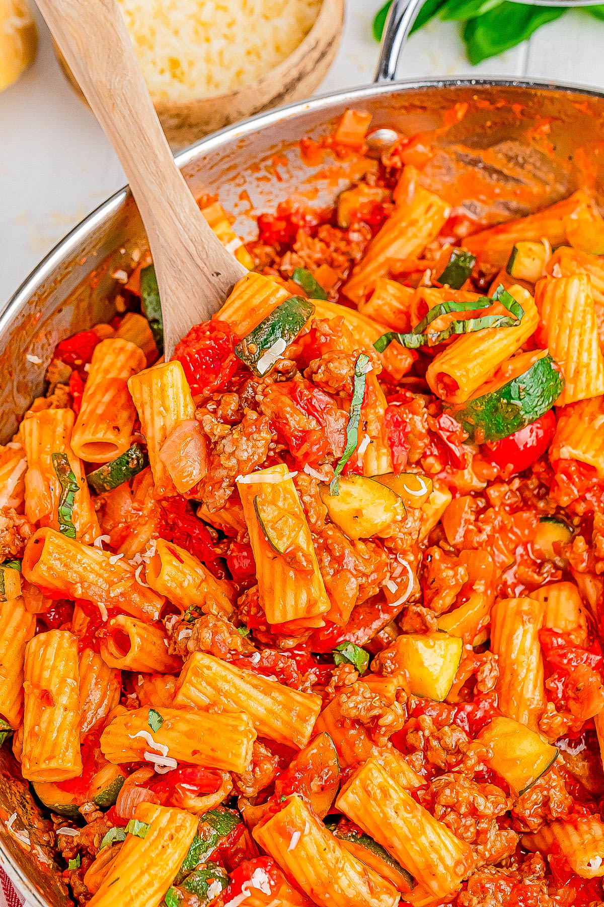 A skillet filled with rigatoni pasta, zucchini, bell peppers, and ground meat in a rich tomato sauce. A wooden spoon is stirring the dish. A cheese grater is visible in the background.