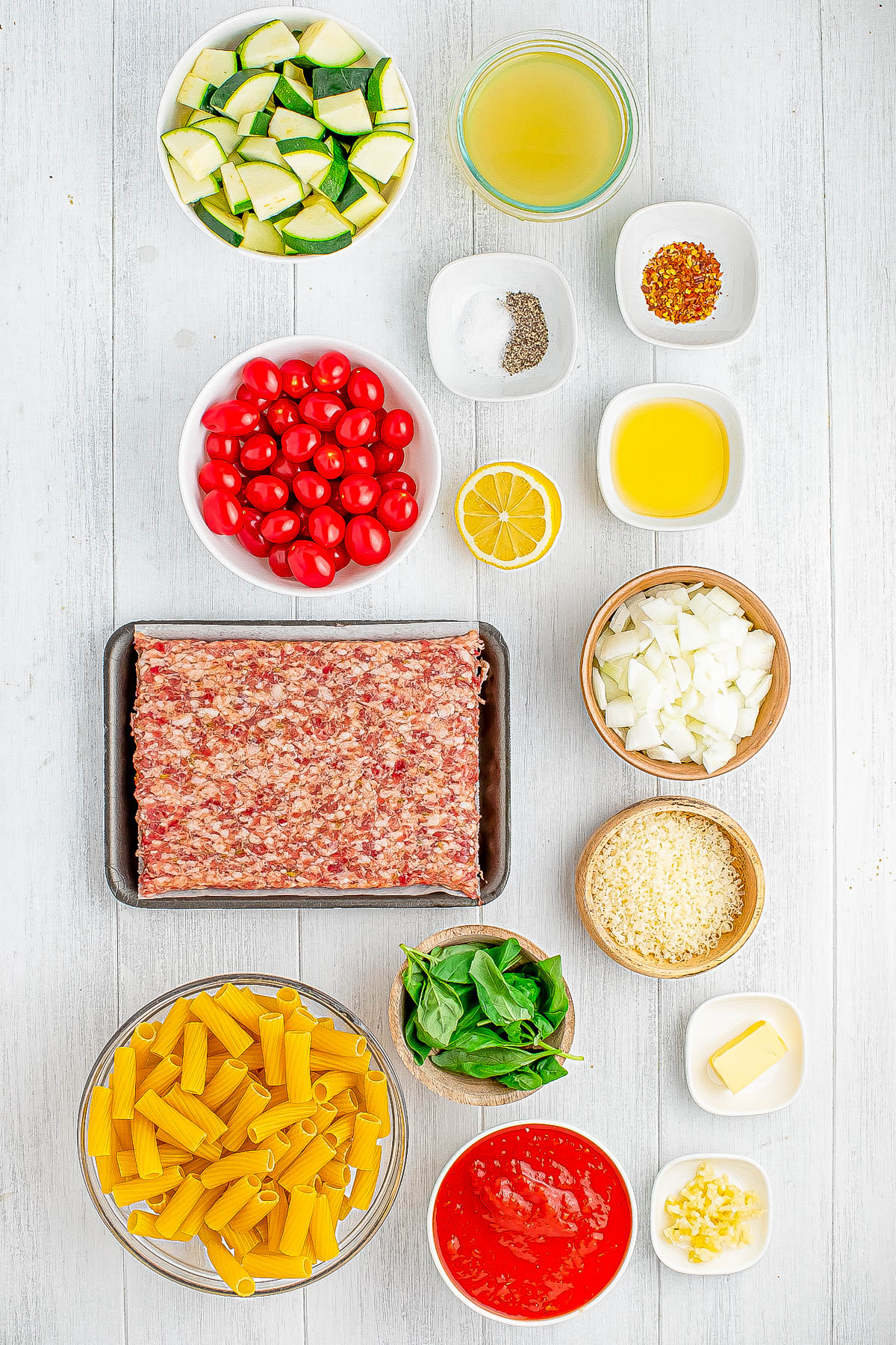 Ingredients arranged on a white surface: zucchini, broth, spices, olive oil, tomatoes, lemon, ground meat, onions, garlic, cheese, penne, spinach, tomato sauce, and butter.